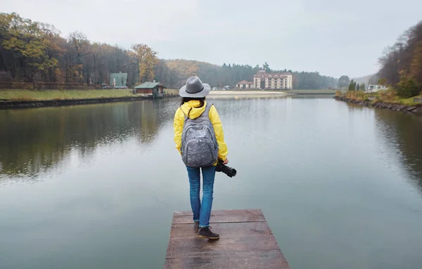 Stylowa młoda kobieta w żółtym wodoodpornym płaszczu i kapeluszu, spacerując po drewnianym molo nad jeziorem w parku jesienią. Widok z tyłu. — Zdjęcie stockowe