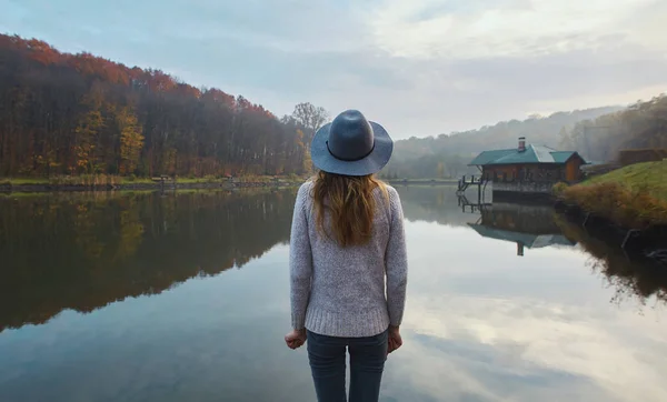 Stilig ung kvinne i varm genser og hatt, som står på brygge ved sjøen i parken om høsten. Sett bakfra . – stockfoto