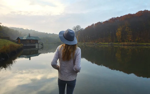 Tyylikäs nuori nainen lämpimässä puserossa ja hatussa, seisoo laiturilla järven rannalla puistossa syksyllä. Taustakuva . — kuvapankkivalokuva
