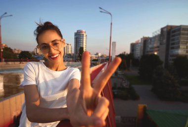 Gözlük, kırmızı etek ve beyaz T-shirt genç güzel kadın Parlak yaz yaşam tarzı portre, el veya Parmaklarla V işareti ile barış işareti gösteren, gün batımında parlak kırmızı köprü üzerinde duran