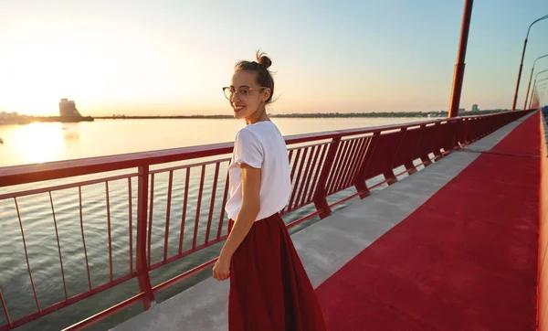 Portrait estival lumineux de jeune jolie femme en lunettes, jupe rouge et T-shirt blanc, debout sur le pont rouge vif, la rivière et le coucher de soleil sur le fond . — Photo