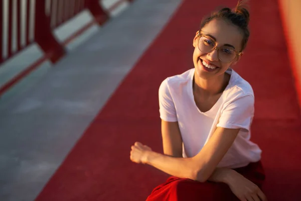 Retrato de cerca de la joven mujer feliz en camiseta blanca, sentada en el puente en las luces del atardecer, mirando a la cámara y sonriendo . — Foto de Stock