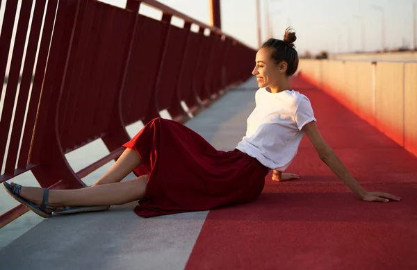 Яркий летний образ жизни молодой красивой женщины в красной юбке и белой футболке, сидящей на ярко-красном полу на мосту . — стоковое фото