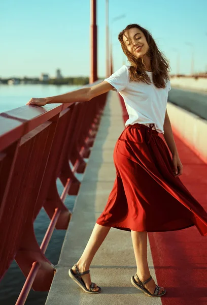 Bright lifestyle portrait of young pretty woman in red skirt and white T-shirt, posing, walking on a bright red floor on the bridge, blowing hair by wind.