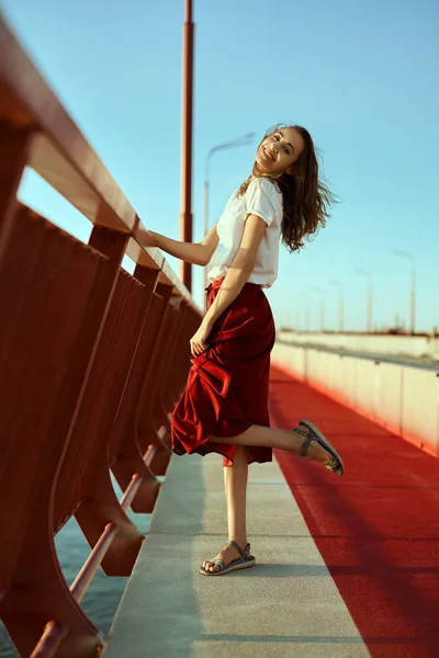 Яркий образ жизни молодой красивой женщины в красной юбке и белой футболке, позирующей, стоящей на ярко-красном полу на мосту, вздувающей волосы от ветра . — стоковое фото