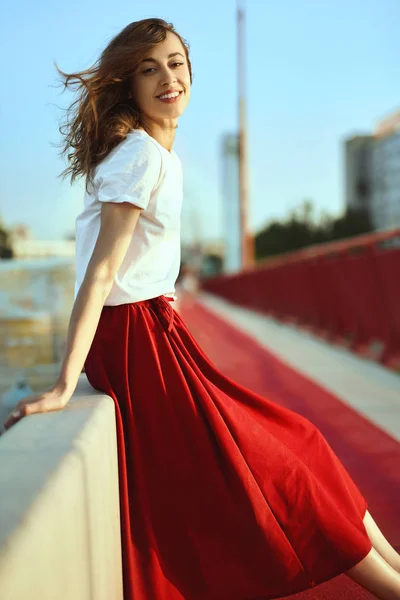 Brillante retrato de estilo de vida de mujer bonita sonriente en falda roja y camiseta blanca, posando, sentada en el parapeto en el puente, soplando pelo por el viento . — Foto de Stock