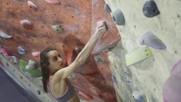 Jovem mulher Rock alpinista está escalando um pedregulho no ginásio de escalada interior. magro bonita mulher fazendo alguns movimentos duros e obter o cume . — Vídeo de Stock