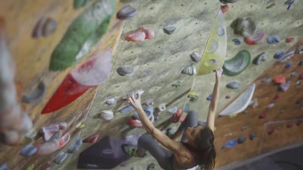 Jovem mulher Rock alpinista está escalando um pedregulho no ginásio de escalada interior. magro bonita mulher fazendo alguns movimentos duros e obter o cume . — Vídeo de Stock