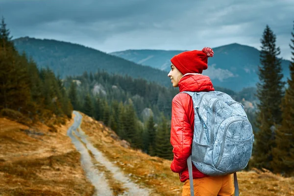 Nő túrázó a hátizsák, visel a piros zakó és narancssárga nadrág, állva a hegyek a háttérben — Stock Fotó