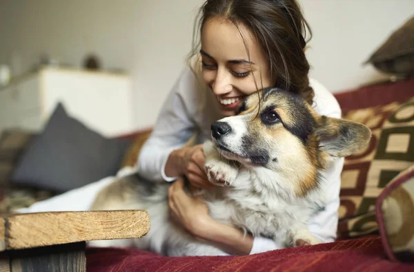 รูปภาพของผู้หญิงที่มีความสุขที่ยิ้มกับสุนัข Welsh Corgi น่ารักของเธอนอนบนโซฟาที่บ้าน . — ภาพถ่ายสต็อก