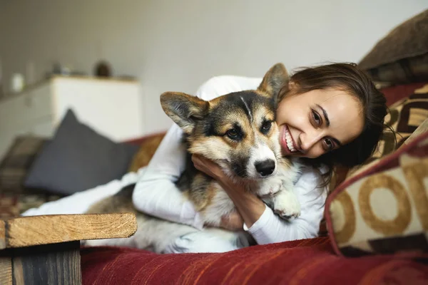 รูปภาพของผู้หญิงที่มีความสุขที่ยิ้มกับสุนัข Welsh Corgi น่ารักของเธอนอนบนโซฟาที่บ้านเพลิดเพลินกับสุดสัปดาห์ตอนเช้า มุ่งไปที่สุนัข . — ภาพถ่ายสต็อก