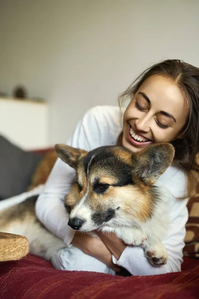 Portrait of happy smiling woman with her cute Welsh Corgi dog lying on couch at home, enjoing morning weekend. Focus on the dog. — Stock Photo, Image
