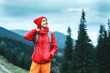 kırmızı ceket ve dağlar arka plan üzerinde duran turuncu pantolon giyen kadın uzun yürüyüşe çıkan kimse ile sırt çantası,