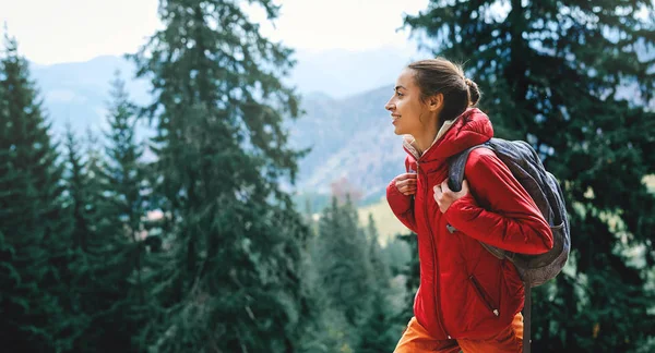 Woman hiker with small backpack wearing in red jacket and orange pants walks by trail through pine woods in the Carpathians mountains — Stock Photo, Image