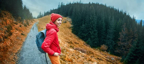 Nő túrázó a hátizsák, visel a piros zakó és narancssárga nadrág, állva a hegyek a háttérben — Stock Fotó