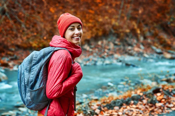 Csinos nő túrázó élénk vörös szabadtéri ruhákat áll a szabadban, a hegyi folyó és az erdő háttér és vidáman nézi kamera — Stock Fotó