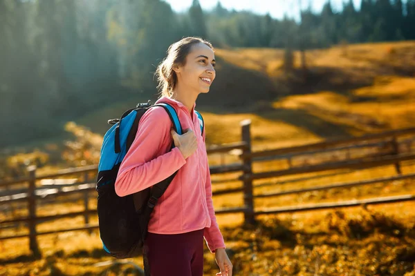 Mujer excursionista con mochila caminando al aire libre. mujer de pie en el prado amarillo y el fondo de bosques verdes en el pueblo de montaña Dzembronya —  Fotos de Stock