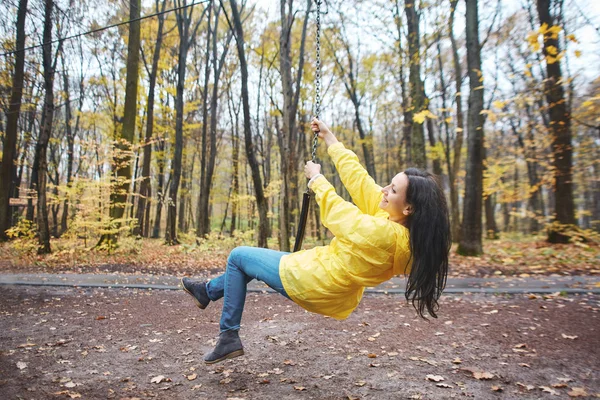 Mujer feliz con chaqueta amarilla en columpio en el parque de otoño. Despreocupada hembra divirtiéndose, disfrutando de un buen día de otoño . — Foto de Stock
