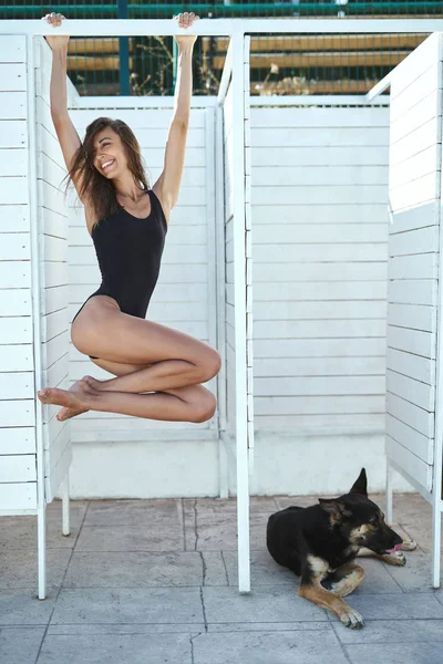 Mujer bronceada atractiva joven con piernas largas en un traje de baño negro posando sobre un fondo blanco. Un gran perro negro está tirado cerca . — Foto de Stock