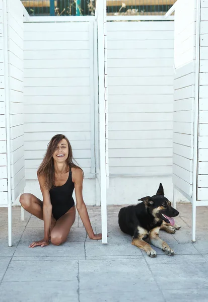 Young attractive tanned woman in a black swimsuit sitting down, posing on a white background. A big black dog is lying nearby. — Stock Photo, Image