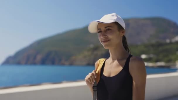 Ritratto esterno di bella donna sportiva sexy elegante in costume da bagno nero e cappuccio bianco passeggiando lungo la baia al mare, contro un mare blu nella giornata di sole poi sorridente alla macchina fotografica . — Video Stock