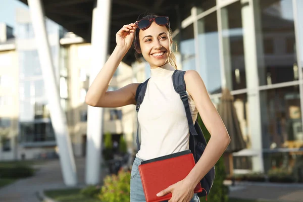 Hermosa mujer feliz en gafas de sol con una mochila que va a la universidad. Joven estudiante universitaria con libros en el campus . — Foto de Stock