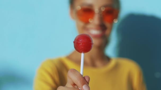 Närbild av ansiktet på en ung garvade skrattande kvinna. glad leende kvinna med röda glasögon stående mot blå bakgrund och glatt slickar en stor röd klubba. — Stockvideo