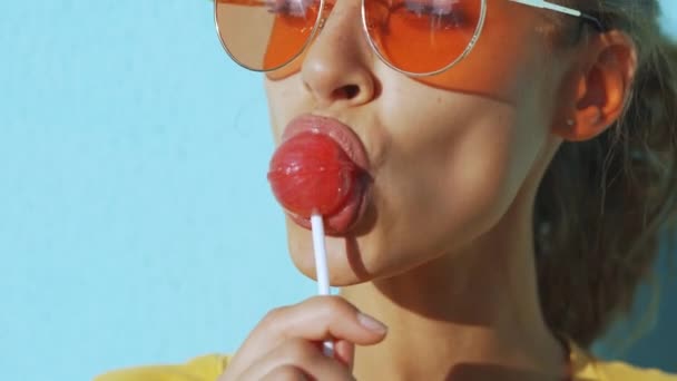 Primer plano de la cara de una joven mujer sonriente bronceada. Mujer alegre con gafas rojas posando sobre fondo azul y lamiendo una gran piruleta roja . — Vídeos de Stock