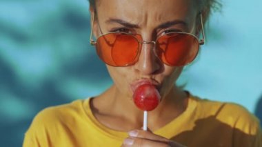 Büyük kırmızı lolipoplu, bronzlaşmış, duygusal bir kadının yüzüne yakın çekim. Kırmızı gözlüklü neşeli kadın mavi arka planda duruyor, lolipop yalıyor ve farklı duygular gösteriyor.