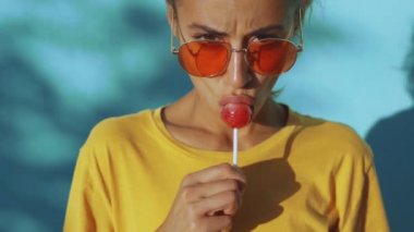 Büyük kırmızı lolipoplu, bronzlaşmış, duygusal bir kadının yüzüne yakın çekim. Kırmızı gözlüklü neşeli kadın mavi arka planda duruyor, lolipop yalıyor ve farklı duygular gösteriyor.