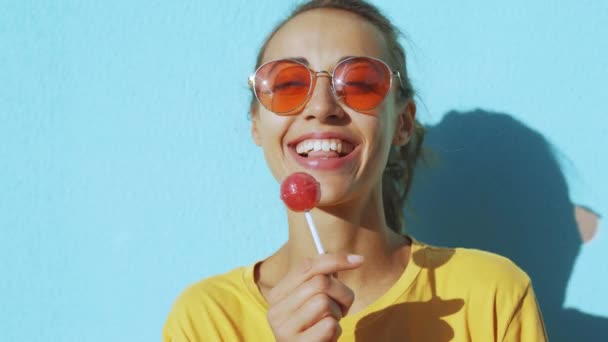 Gros plan du visage d'une jeune femme rieuse bronzée. femme souriante joyeuse avec des lunettes rouges debout sur fond bleu et léchant joyeusement une grosse sucette rouge . — Video