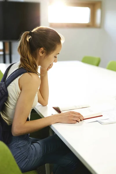 Cheerful young female student of IT school sitting at table in classroom, reading notes and information for copybook Royalty Free Stock Images