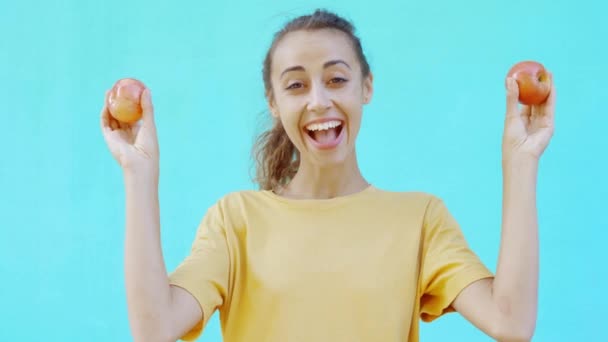 Menina muito sorridente está posando em um fundo ciano brilhante colorido, e mostra maçãs amarelas-vermelhas maduras frescas saborosas. mulher mostra felicidade, prazer e emoções positivas — Vídeo de Stock