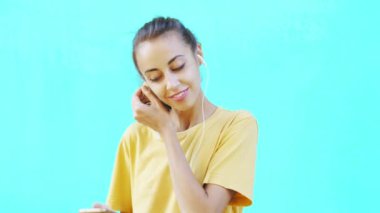 Akıllı telefon kullanan ve kulaklıkla müzik dinleyen güzel, gülümseyen genç bir kadın renkli parlak bir siyan arka planda poz veriyor. Kadınlar zevk, keyif ve olumlu duygular gösterir..