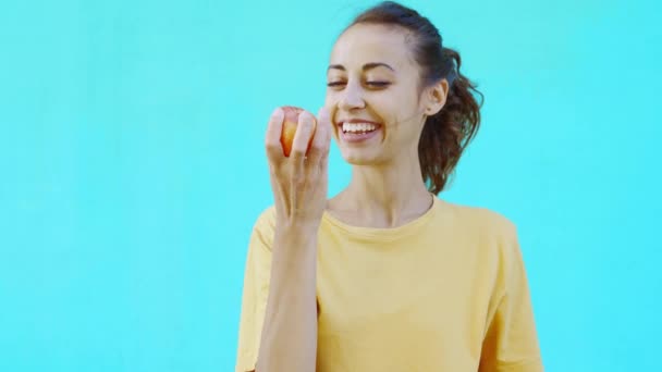 Pretty smiling girl eats fresh ripe tasty yellow-red apple, posing on a colorful bright cyan background. — Stock Video