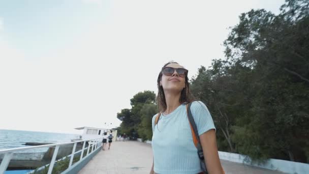 Portrait extérieur de belle jeune femme souriante gaie en tenue décontractée et petit sac à dos debout au bord de la mer, contre une mer bleue . — Video