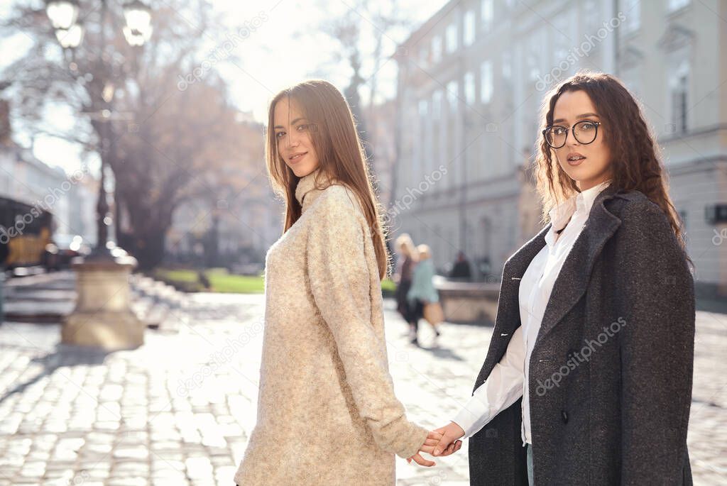 Smiling young lesbian couple holding hands, walking in the street of the city at sunny autumn day.