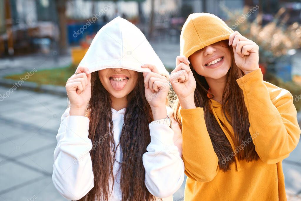 Portrait of funny girl friends in colored hoodies posing in hood with smile on the street at autumn day.