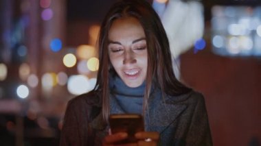 Güzel, gülümseyen bir kadının gece vakti akıllı telefon kullanarak yürüdüğü yakın plan.