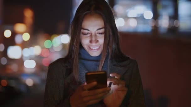 Nahaufnahme einer lächelnden Frau mit Smartphone, die nachts in der Stadt spazieren geht — Stockvideo