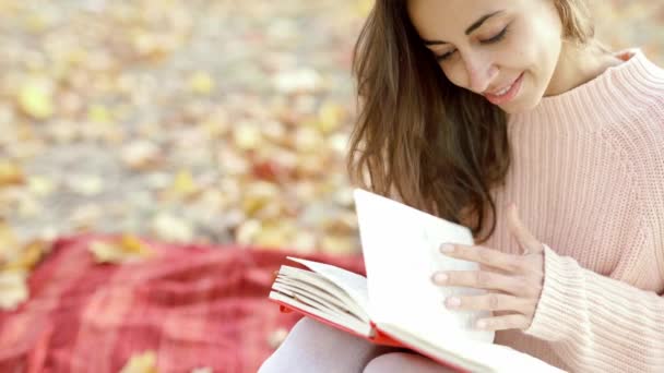 Mujer atractiva joven en suéter de punto rosa sentado en la manta roja en el bosque de otoño, leyendo un cuaderno . — Vídeo de stock