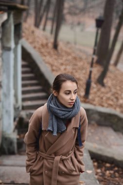 Sonbahar parkında bej palto ve sıcak örgü eşarp giyerek yürüyen güzel bir kadının sonbahar portresi..
