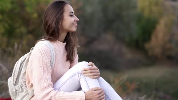 Closeup of pretty young girl in knited pink sweater with small backpack sitting on the blanket in the autamn forest and smiling. — Stock Video