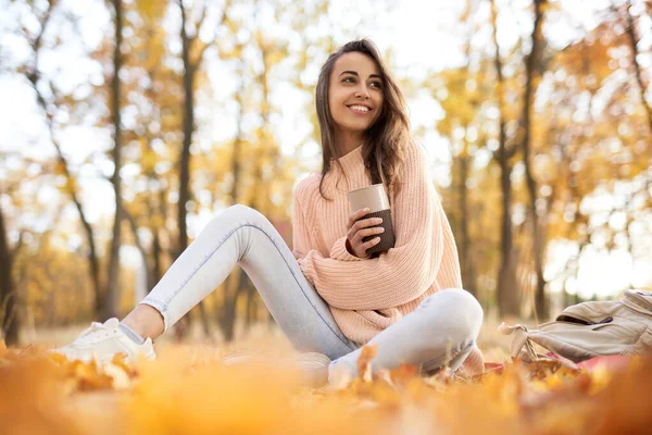 Söpö hymyilevä nainen syyspuistossa, istuu peiton päällä, juo kahvia, nauttii lämpimästä aurinkoisesta säästä. Syksyn käsite . — kuvapankkivalokuva