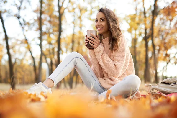 Søt, smilende kvinne i høstparken, sitter på et teppe, drikker kaffe, nyter varmt, solrikt vær. Fall-konsept . – stockfoto