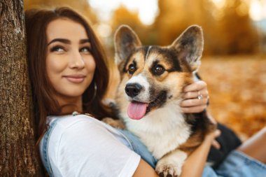 Galli Corgi Pembroke köpeğiyle birlikte parkta gülümseyen mutlu kadın. Genç bayan hayvan sahibi. Sonbaharda parkta büyük bir hayvan. Turuncu yeşillikte..