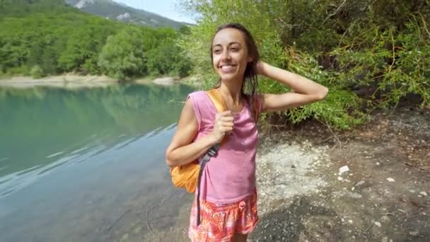 Happy smiling young slender woman with small orange backpack walking along mountain lake with clear turquoise water — Stock Video