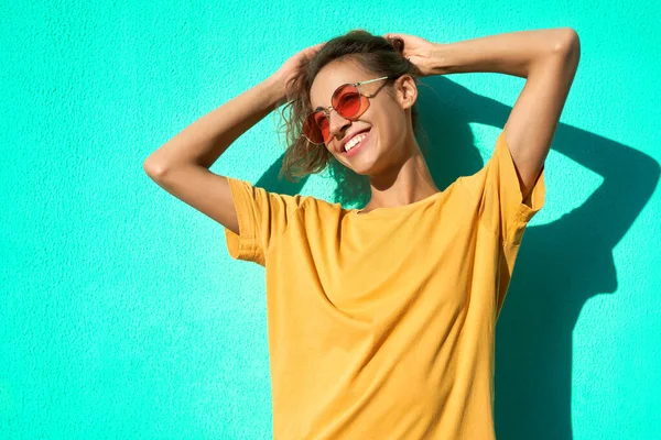 Alla moda allegra giovane donna in posa su sfondo blu, indossando giallo t-short e occhiali rossi alla moda . — Foto Stock