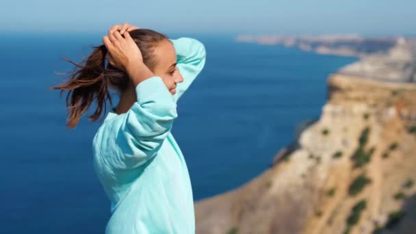 Porträt einer glücklichen jungen Frau, die auf einer Klippe vor atemberaubendem Meer und Himmel steht und die Haare im Wind wehen — Stockvideo