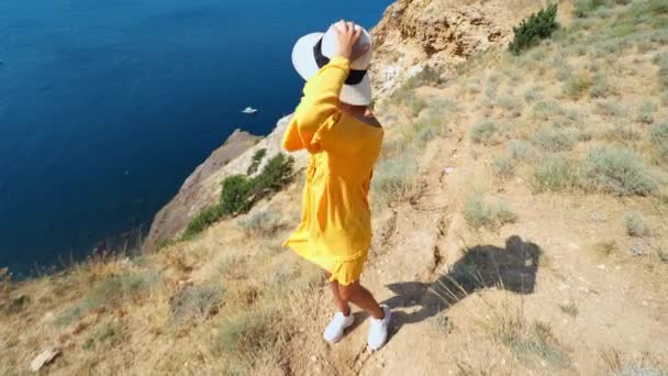 Happy smiling woman in bright yellow dress and hat with backpack standing on cliff edge — Stock Video